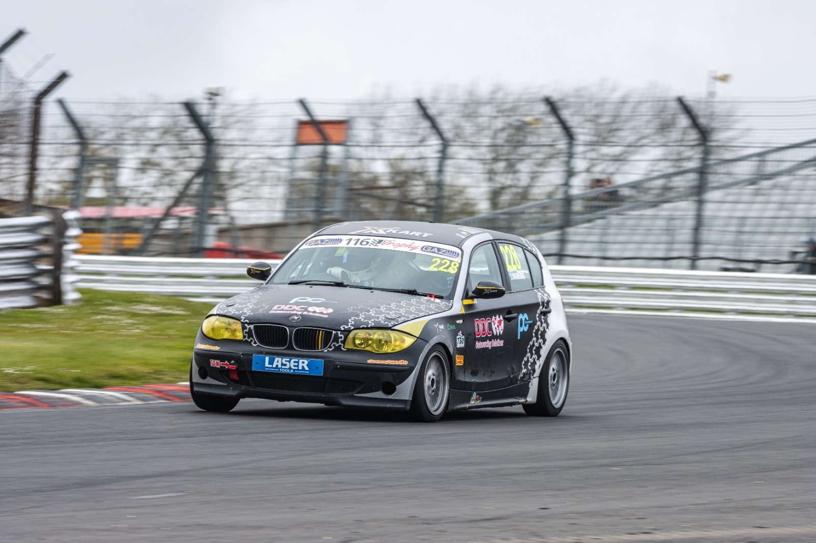 bmw 116 trophy of james wareing on race circuit with metal fencing in background grass and red and white curbing on apex